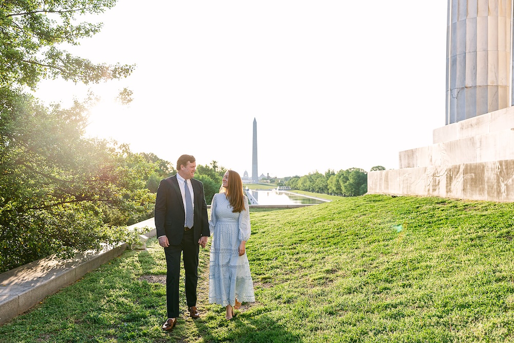 Couple walks on lawn at Lincoln Memorial with Washington Monument behind them. Spring wedding engagement photo session. Sarah Bradshaw Photography.