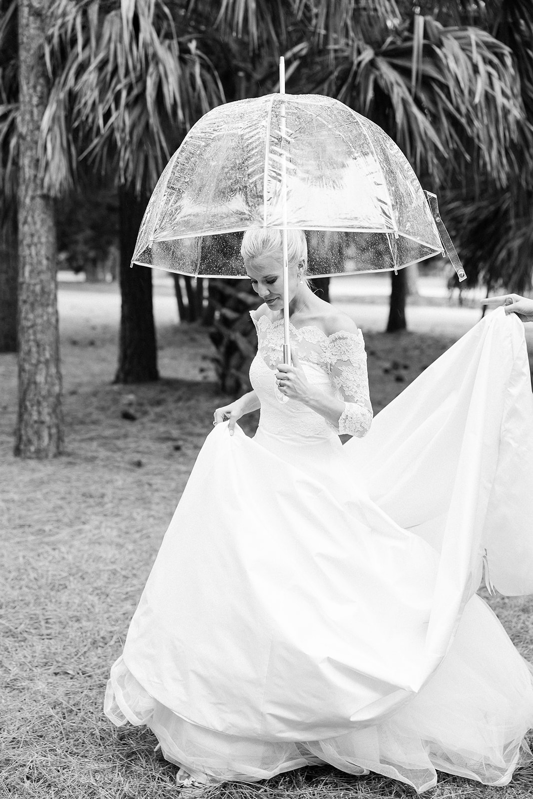 Black and white portrait of bride walking with clear umbrella, holding up wedding dress. Classic Black Tie wedding in Georgetown, SC. Sarah Bradshaw Photography