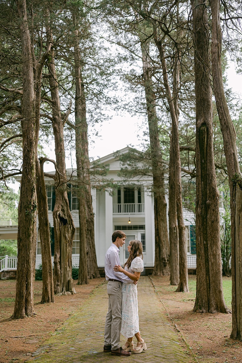 couple poses in front of historic home and trees. southern wedding engagement session, sarah bradshaw photography.