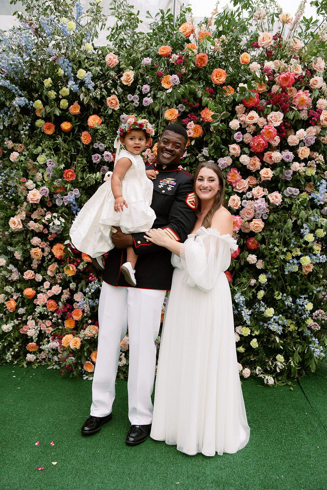 bride in long sleeves wedding dress and groom in military uniform pose with flower girl daughter in front of floral wall. Spring, private home tented wedding in south carolina. Sarah Bradshaw Photography