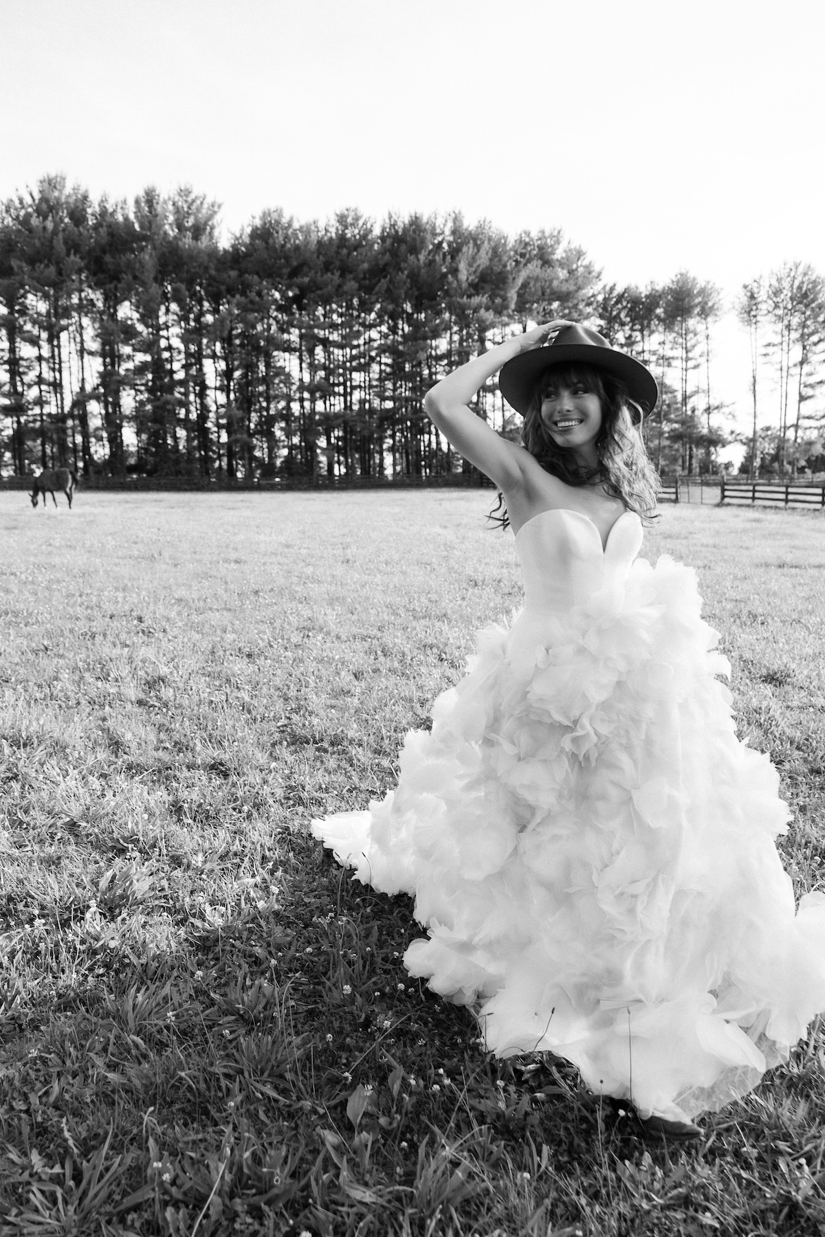 black and white photo of bride in strapless wedding dress with ruffle skirt walking in field with hat. chic equestrian wedding inspiration at Red Fox Inn, Middleburg Virginia. Sarah Bradshaw