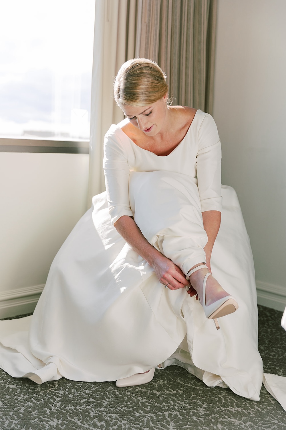 bride in simple no lace wedding dress with scoop neckline and three quarters sleeves, putting on bridal heels. Classic timeless winter wedding, woodlawn manor, alexandria va. Sarah bradshaw photography